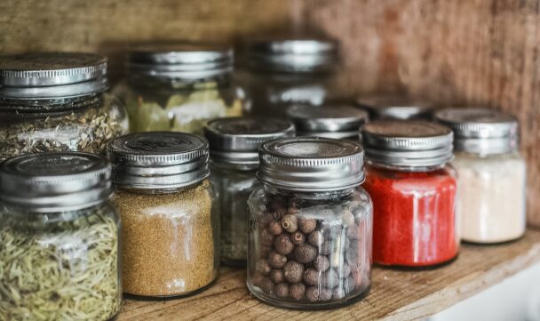 Glass spice jars filled with colorful ground and whole spices like turmeric, peppercorns, and paprika on a rustic wooden shelf.