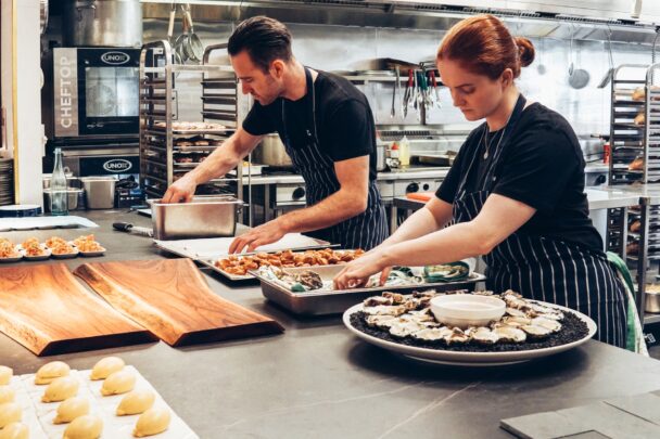 Two beginner chefs preparing food in a professional kitchen, showcasing essential cooking techniques and teamwork as part of learning the basics of cooking.
