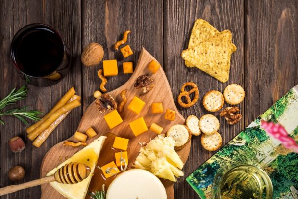 Top view of assorted cheeses, crackers, pretzels, and wine on a wooden board—featuring cheddar cubes, soft brie, nuts, and gourmet snacks