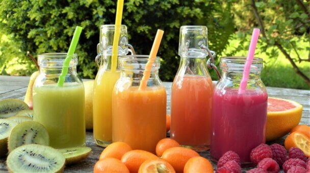 Five colorful smoothies in glass jars with straws, surrounded by fresh fruits like kiwi, raspberries, oranges, and grapefruit on a wooden table outdoors