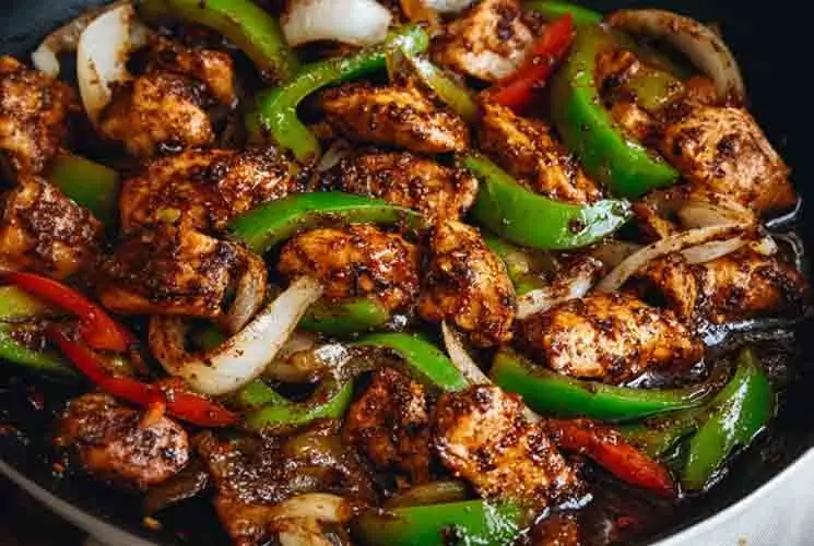 Close-up of stir-fried black pepper chicken with bell peppers and onions in a glossy pepper sauce, cooked in a skillet.