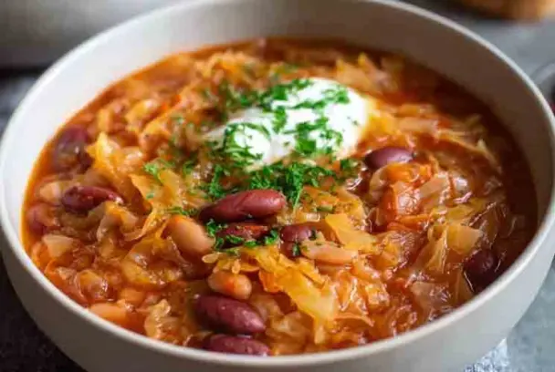 Close-up of a traditional Croatian cabbage and bean stew (Zelje i Grah) featuring red beans, sauerkraut, and sour cream, garnished with fresh parsley in a white bowl.