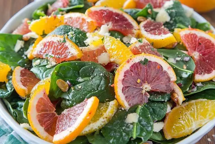 Close-up of a spinach salad topped with sliced oranges, grapefruit, avocado, toasted nuts, and grated cheese in a white bowl on a wooden surface.