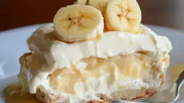A slice of banana pudding on a white plate, showing layers of vanilla pudding, whipped cream, banana slices, and a cookie crust, with a fork beside it.