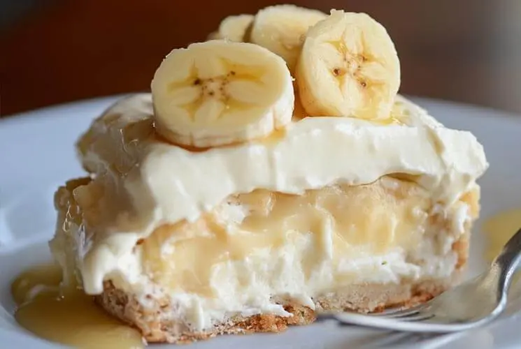 A slice of banana pudding on a white plate, showing layers of vanilla pudding, whipped cream, banana slices, and a cookie crust, with a fork beside it.