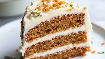 Close-up of a moist, triple-layer carrot cakes with creamy white frosting and crumbled topping, garnished with herbs on a white plate.
