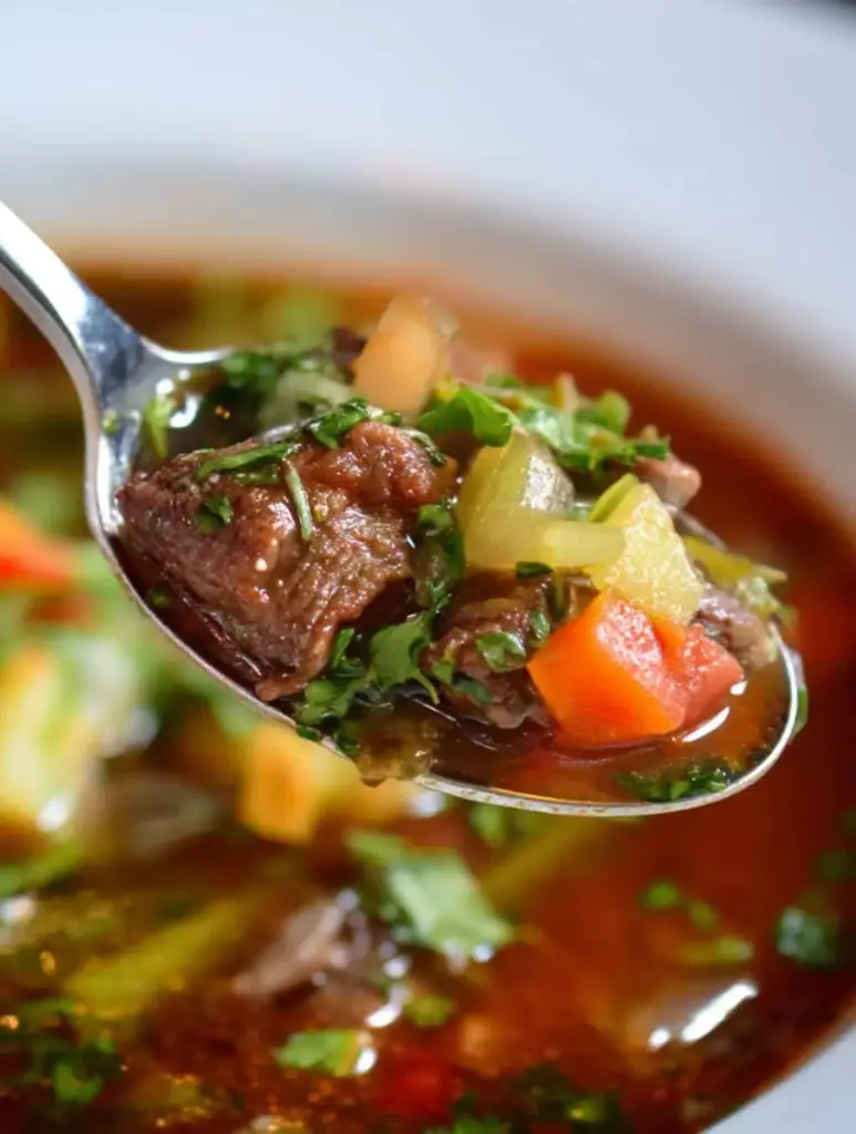 Close-up of a spoonful of vegetable beef soup featuring tender beef chunks, diced carrots, celery, and herbs above a bowl of savory broth-based soup.
