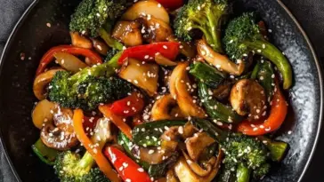 Top-down view of a vegetable stir fry in a black bowl, featuring broccoli, red bell peppers, mushrooms, carrots, and green onions, sprinkled with sesame seeds.