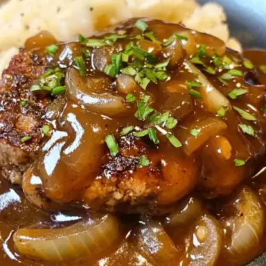 Close-up of a juicy hamburger steak covered in thick brown onion gravy, garnished with chopped fresh herbs, served alongside creamy mashed potatoes in a dark bowl.