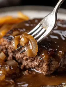 Close-up of a fork lifting caramelized onions and gravy from a juicy hamburger steak, revealing the tender inside of the meat and rich brown sauce.