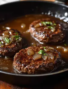 Three seared hamburger steaks simmering in a skillet filled with brown onion gravy, topped with chopped onions and fresh herbs.
