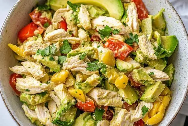 Top view of a bowl filled with avocado chicken salad featuring shredded chicken, chopped avocado, yellow bell pepper, cherry tomatoes, and cilantro.