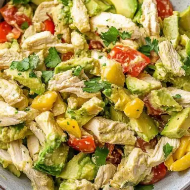 Close-up of a vibrant avocado chicken salad with shredded chicken, diced avocado, cherry tomatoes, mango, and cilantro in a ceramic bowl.
