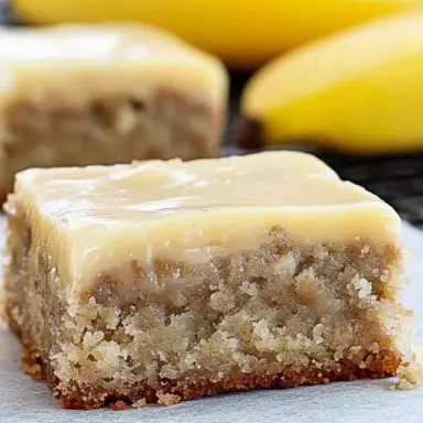Close-up of a thick, moist banana bread brownie topped with glossy brown butter frosting, with fresh bananas blurred in the background.