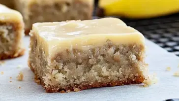 A close-up of a thick banana bread brownie topped with smooth brown butter frosting, resting on parchment paper with bananas in the background.