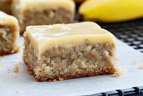 A close-up of a thick banana bread brownie topped with smooth brown butter frosting, resting on parchment paper with bananas in the background.