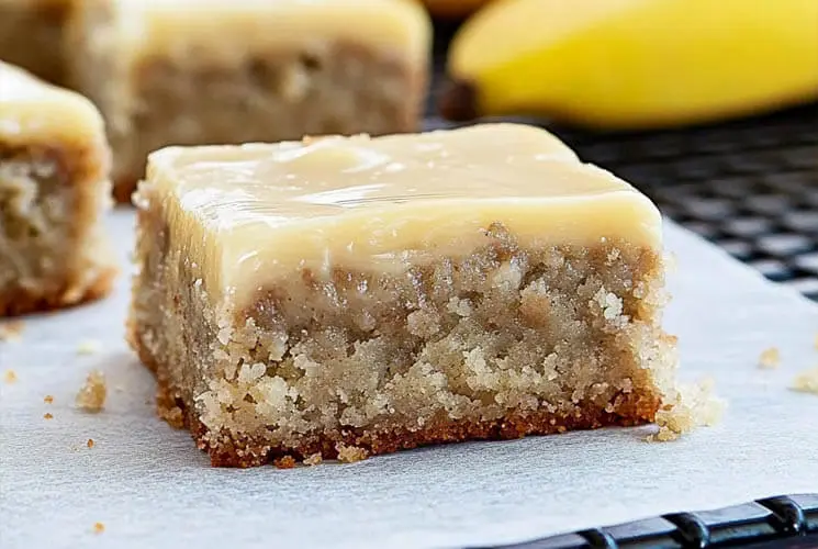 A close-up of a thick banana bread brownie topped with smooth brown butter frosting, resting on parchment paper with bananas in the background.
