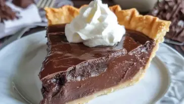 A close-up of a slice of creamy chocolate pie with a golden flaky crust and a swirl of whipped cream on top, served on a white plate.