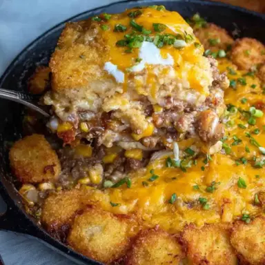 A close-up of cheesy cowboy casserole with ground beef, corn, and golden tater tots in a cast iron skillet, topped with sour cream and chopped chives.