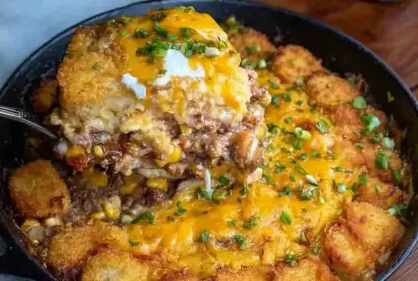 Cast iron skillet filled with cowboy casserole made of ground beef, corn, cheese, and tater tots, with a cheesy portion lifted by a serving spoon.