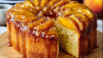 Close-up of a Peach Cobbler Pound Cake on a wooden board, topped with glossy caramelized peach slices and rich brown sugar glaze, with a slice cut out.