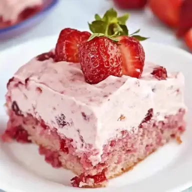 Close-up of a slice of moist strawberry sheet cake topped with fluffy pink strawberry frosting and fresh strawberries on a white plate.