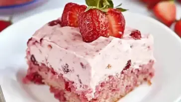 A slice of moist strawberry sheet cake with pink strawberry frosting, topped with halved fresh strawberries, served on a white plate with whole strawberries in the background.