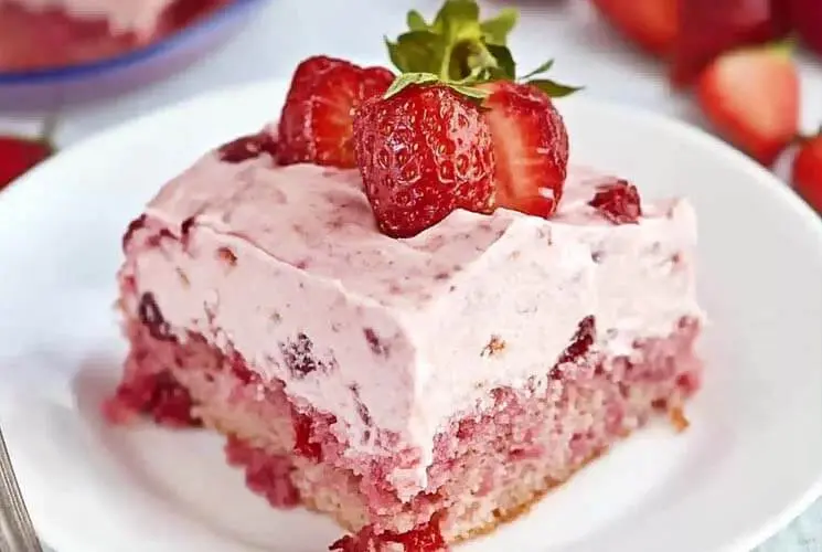 A slice of moist strawberry sheet cake with pink strawberry frosting, topped with halved fresh strawberries, served on a white plate with whole strawberries in the background.