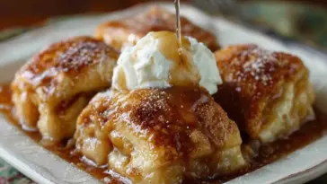 Close-up of golden-brown Amish Apple Dumplings topped with whipped cream and drizzled with cinnamon syrup on a white plate.