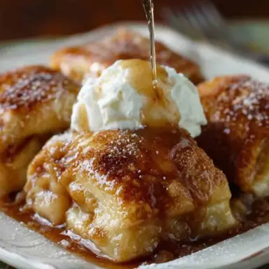 Close-up of golden-brown Amish Apple Dumplings topped with whipped cream and drizzled with cinnamon syrup on a white plate.