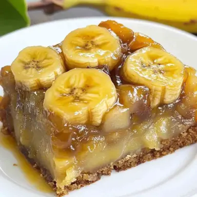 A thick slice of banana upside down cake with glossy caramelized banana slices on top, served on a white plate with fresh bananas in the background.