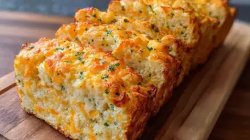 Sliced loaf of golden-baked Cheddar Cheese Quick Bread with parsley and melted cheese on a wooden cutting board.