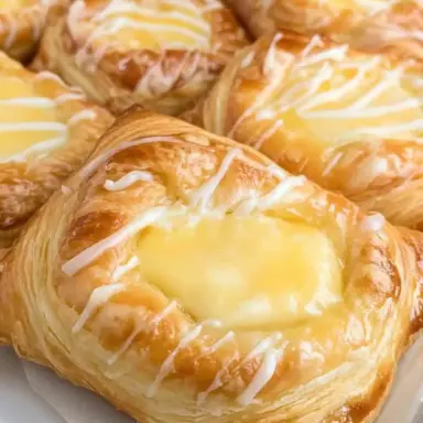 Close-up of golden, flaky Cheese Danishes filled with sweet cream cheese and drizzled with vanilla glaze on a parchment-lined tray.