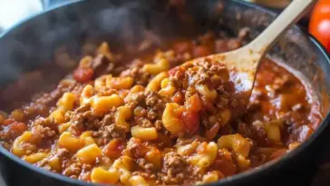 Steaming pot of cheesy American goulash with ground beef, elbow macaroni, and tomato sauce being stirred with a wooden spoon