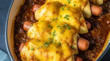 Baked Chili Cheese Dog Bake with crescent-wrapped hot dogs, topped with melted cheddar cheese in a rich chili base, served in a white casserole dish.