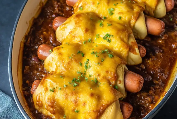 Baked Chili Cheese Dog Bake with crescent-wrapped hot dogs, topped with melted cheddar cheese in a rich chili base, served in a white casserole dish.