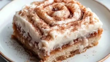 A thick slice of cinnamon roll cake topped with creamy glaze and cinnamon sugar, showing its moist layers and cinnamon swirl on a white plate.