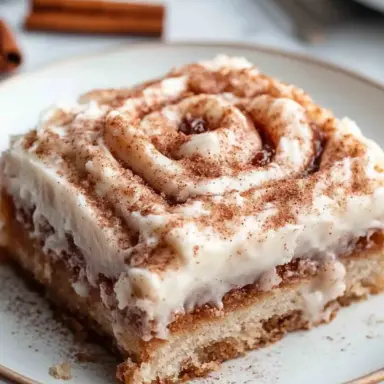 A close-up of a slice of cinnamon roll cake on a white plate, showing the gooey cinnamon swirl and creamy glaze, topped with a dusting of cinnamon.