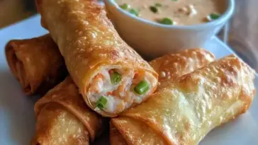 Stack of crispy crab rangoon egg rolls filled with cream cheese, crab, and green onions, served with a creamy dipping sauce in a white bowl.