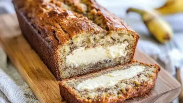 A loaf of cream cheese filled banana bread on a wooden board, sliced to reveal a thick, creamy filling inside a moist, golden banana bread.