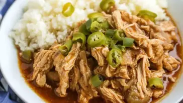 Plate of shredded Crock Pot Mississippi Chicken topped with sliced pepperoncini peppers, served with white rice and rich, savory sauce