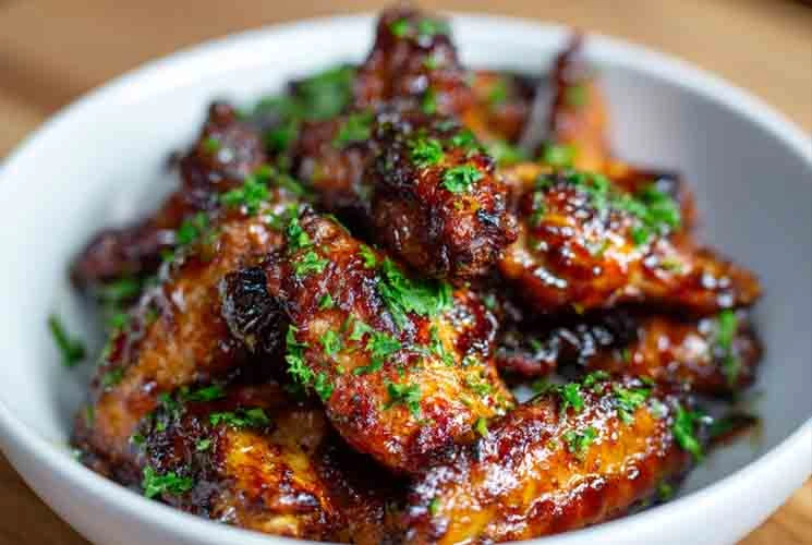Close-up of sticky, glazed Crockpot BBQ chicken wings in a white bowl, topped with fresh chopped parsley and glistening with barbecue sauce.