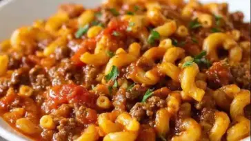 Hearty American goulash with elbow macaroni, ground beef, and tomato sauce served in a white bowl and garnished with chopped parsley.