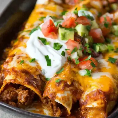 Close-up of cheesy ground beef enchiladas topped with sour cream, diced avocado, tomatoes, and fresh cilantro in a black serving dish.
