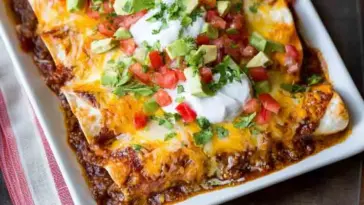 Baked ground beef enchiladas in a white dish, topped with melted cheese, sour cream, diced tomatoes, avocado, and chopped cilantro.