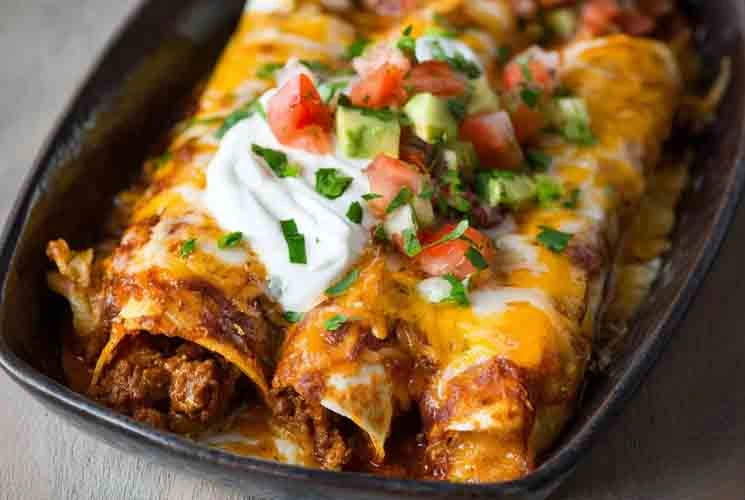 Cheesy ground beef enchiladas in a dark baking dish topped with sour cream, diced avocado, tomatoes, and chopped cilantro.