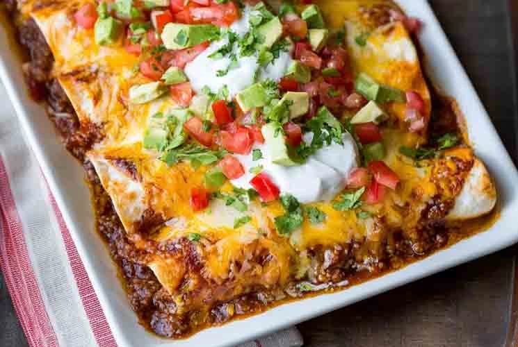 Baked ground beef enchiladas in a white dish, topped with melted cheese, sour cream, diced tomatoes, avocado, and chopped cilantro.