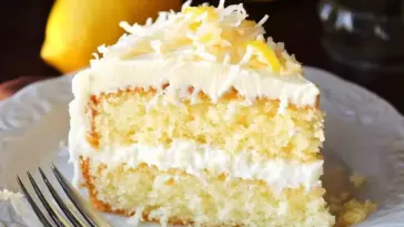 Slice of moist lemon and coconut cake with creamy white frosting and shredded coconut topping, served on a white plate with a fork.