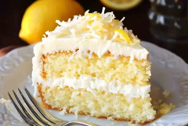 Slice of moist lemon and coconut cake with creamy white frosting and shredded coconut topping, served on a white plate with a fork.