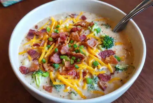 Creamy loaded broccoli and potato soup topped with melted cheddar, bacon pieces, and fresh chopped green onions in a white bowl with spoon.